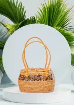 Vue de face du sac à main en raphia tressé jaune soleil avec anses en cuir et médaillons en bois, fond de feuilles de palmier