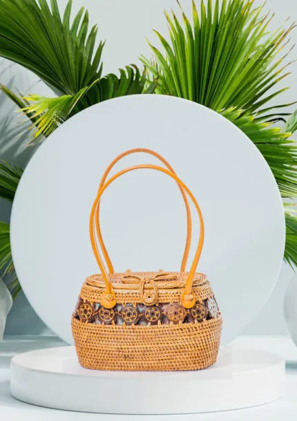 Vue de face du sac à main en raphia tressé jaune soleil avec anses en cuir et médaillons en bois, fond de feuilles de palmier