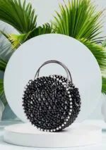 Vue de face du sac à main rond noir à perles facettées avec liseré de perles blanches, fond de feuilles de palmier