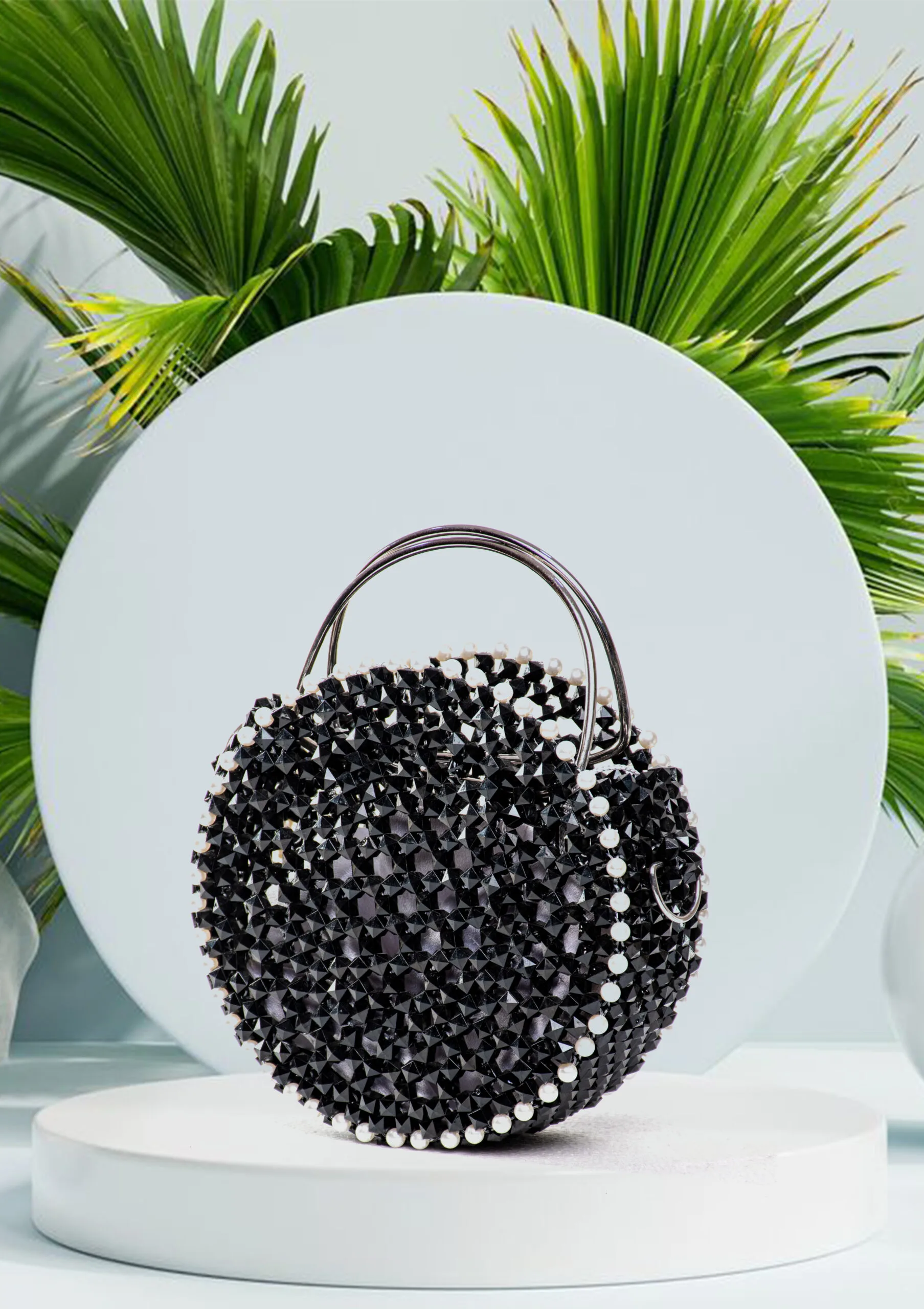 2 (1) Vue de face du sac à main rond noir à perles facettées avec liseré de perles blanches, fond de feuilles de palmier
