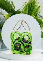 Vue de face du sac à main tressé en disques vert fluo avec tissu violet visible et anses anneaux dorés, fond de feuilles de palmier