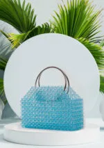 Vue de face du sac à main en perles bleu azur transparentes à anses anneaux chromés, fond de feuilles de palmier