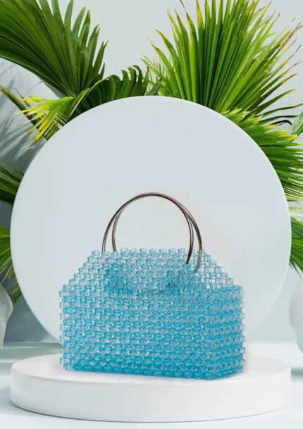 Vue de face du sac à main en perles bleu azur transparentes à anses anneaux chromés, fond de feuilles de palmier