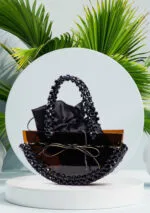 Vue de face du sac à main noir à perles facettées avec col en perles ambre translucide et anse tressée, fond de feuilles de palmier