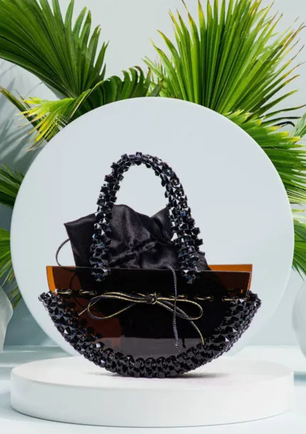 Vue de face du sac à main noir à perles facettées avec col en perles ambre translucide et anse tressée, fond de feuilles de palmier
