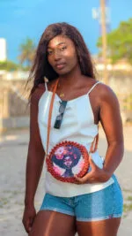 Mannequin portant en bandoulière le sac à main rond perlé rouge bordeaux à motif visage fleuri, sur une plage