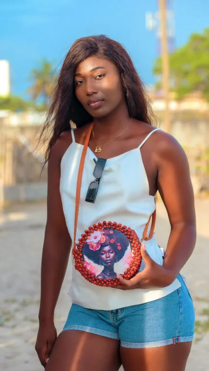 Mannequin portant en bandoulière le sac à main rond perlé rouge bordeaux à motif visage fleuri, sur une plage