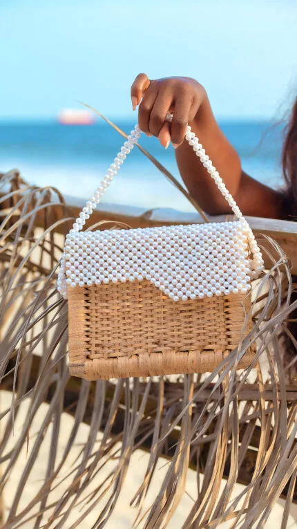 Gros plan sur le sac à main en rattan tressé à perles blanches, suspendu à la main face à la mer
