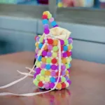 Vue de face du mini sac seau perlé multicolore à doublure satin crème, posé sur une table en bois